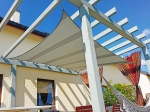 Beige premium waterproof sun sail 3x4 m mounted on a wooden terrace pergola, protecting against sun and rain.