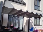 Grå horisontell premium Decor hissgardin måttbeställd och monterad över terrassen vid ett hus med trädgård.