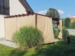 Οριζόντια premium ρωμαϊκή κουρτίνα Decor διαστάσεων 2,5x3 μ., τοποθετημένη κάτω από καφέ πέργκολα στον κήπο.