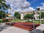 Béžová slnečná clona HDPE 4,2x4,2x5,9 na oceľových stĺpoch nad drevenou terasou pri detskom ihrisku.
