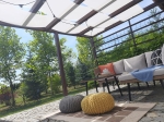 Világos napellenző sáv pergola szerkezeten, amely kerti bútorokat, puffokat és szőnyeget árnyékol a teraszon.