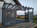 Graphitgraue Pergola mit weißen Sonnenschutzlamellen auf einer gepflasterten Terrasse am Haus.