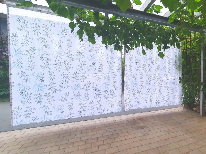Skreddersydd nettinggardin med et delikat botanisk bladmotiv, opphengt under en terrassepergola.