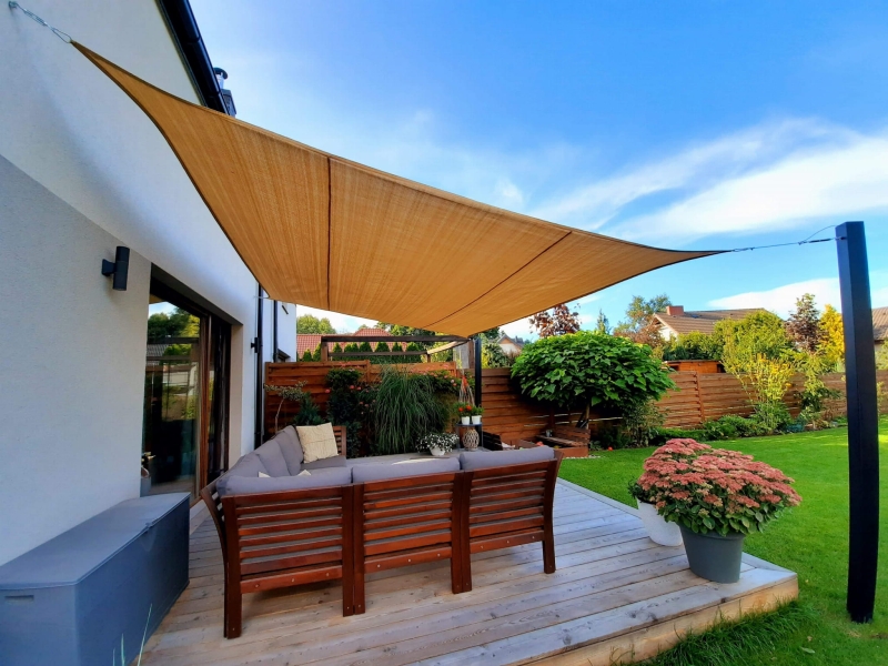 Toldo vela de HDPE beige a medida sombreando una terraza con muebles de exterior y jardín verde.
