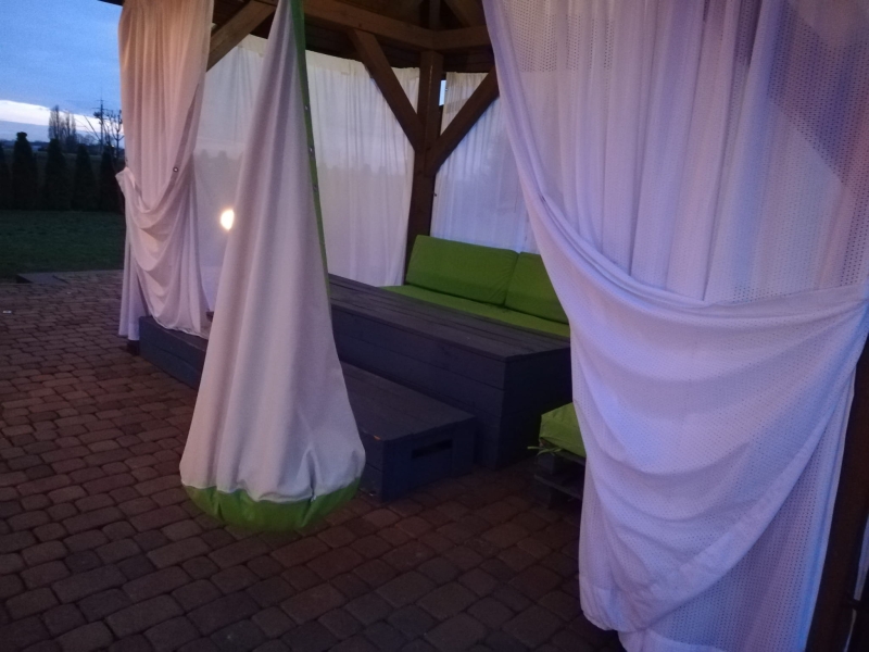 Maßgefertigte Netzgardine im hölzernen Gartenpavillon mit grünen Möbeln.