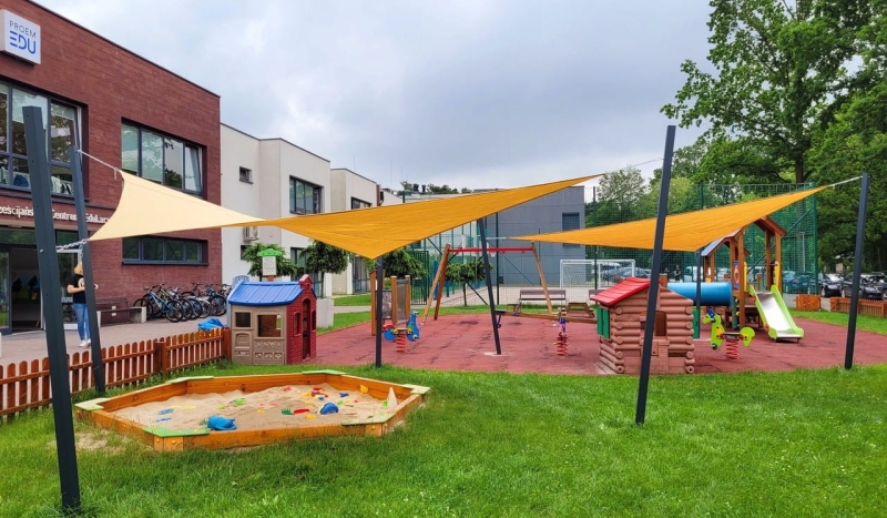 Modern bir anaokulu binasının yanındaki çocuk oyun alanının üzerine monte edilmiş özel yapım sarı güneş yelkenleri.