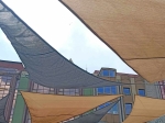 Grafitová a piesková trojuholníková slnečná clona HDPE 6x6x6 namontovaná nad terasou na pozadí budov.
