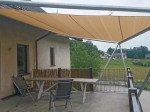Toldo vela cuadrado beige HDPE 5x5 instalado sobre una terraza con muebles de jardín y barandilla.