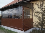 Prozorna terasna zavesa po meri z rjavim spodnjim robom, nameščena na konstrukcijo nadkrite terase.