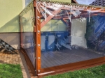 Skræddersyet gennemsigtig terrasseafdækning med brun kant, monteret på en træpavillon med havemøbler.