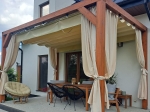 Beige horizontal PVC Roman blind made to measure mounted on a wooden terrace pergola with curtains and furniture.