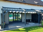 Store romain horizontal en PVC sur mesure installé sur une pergola de terrasse en graphite près d'une maison moderne.
