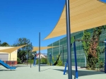 Vela ombreggiante in HDPE 3x3x4,2 che ombreggia un moderno parco giochi con strutture blu.