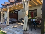 Cortina de cenador Premium en color beige montada en una pérgola de madera con muebles de jardín.