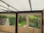 Custom-made transparent terrace cover with beige border, mounted under a roof with a view of the garden.