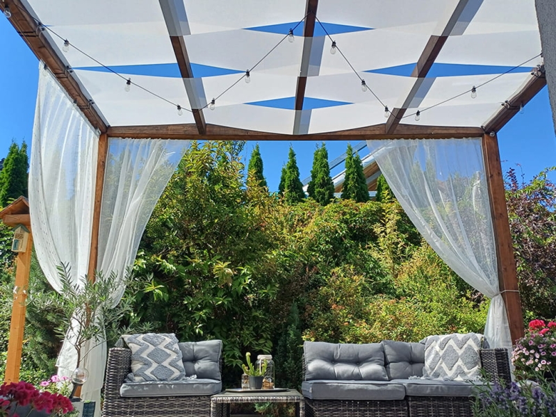 Vit solskyddsskugga för paviljong monterad på en träpergola med belysning i en trädgård med konstrottingmöbler.