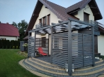 Toldo tipo banda gris moderno instalado en una pérgola de madera junto a la terraza de la casa.