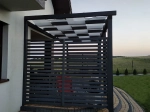 Tamna drvena pergola s krovom isprepletenim bijelim trakama za zaštitu od sunca na terasi uz kuću.