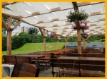 Witte schaduwstrook gemonteerd op een houten pergola in een restauranttuin met bankjes en bloemen.