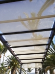 Custom-made white Roman sun shade strip mounted on a brown terrace pergola surrounded by palms.