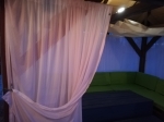 Фіранка під замовлення, підвішена в садовій альтанці, що закриває диван із зеленими подушками.