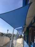 Toldo vela de jardín azul impermeable a medida instalado sobre un camino junto a una playa de arena y un edificio.