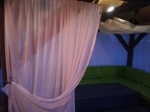 Perdea roz pe terasă, care maschează un colț de relaxare cu o canapea verde într-un foișor de grădină.