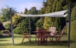 Vela ombreggiante bianca 3,2x3,2 m tesa sopra un set di mobili da giardino in legno in un giardino soleggiato.