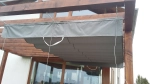 Γκρι οριζόντια ρωμαϊκή κουρτίνα premium Decor 3x4 τοποθετημένη κάτω από ξύλινη κατασκευή πέργκολας στη βεράντα.