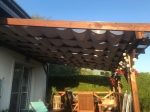 Μπεζ οριζόντια ρωμαϊκή κουρτίνα Decor 3x4 κάτω από τη στέγη ξύλινης πέργκολας στον κήπο.
