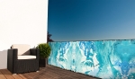 Balkonscherm met blauw abstract patroon, gemonteerd aan de terrasreling naast een tuinstoel.