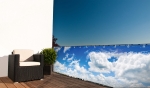 Balkon met donkere fauteuil en groene plant, afgeschermd door een balkonzeil met een print van blauwe lucht en witte wolken.