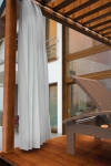 Custom-made net curtain hanging on a terrace overlooking a modern house and a wooden sun lounger.