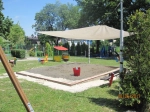 Vela de sombra branca 3,2x2,5 m montada sobre uma caixa de areia num parque infantil, proporcionando sombra num dia de sol.