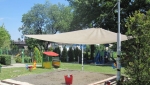 Bijelo kvadratno sjenilo za sunce 3,2x3,2 m montirano iznad pješčanika na dječjem igralištu.