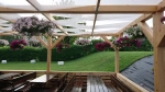 Wit zonnezeil voor overkapping 1x5 m gemonteerd op een houten pergola met hangbloemen en banken.