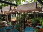 Μπεζ οριζόντια ρωμαϊκή κουρτίνα premium Decor 3x4 που καλύπτει τραπέζια σε κήπο εστιατορίου ανάμεσα σε φυτά.