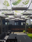 Bande d'ombrage graphique motif feuilles pour tonnelle, montée sur une pergola en bois au-dessus d'une terrasse meublée.