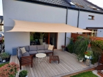 Beige HDPE schaduwdoek 4x4m gespannen over een terras met grijze tuinmeubelen en een lichte gevel.