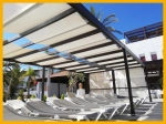 Store romain pare-soleil clair 2,1x5 m monté sur une pergola marron au-dessus de transats près de la piscine de l'hôtel.