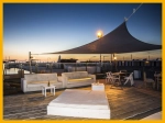 Toldo vela impermeable premium gris beige 4,2x4,2x5,9 cubriendo una terraza con sofás junto al mar al atardecer.