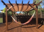Bruine pergola op terras met een beige schuifzeil op kabels, dat een hangmat tegen de zon beschermt.