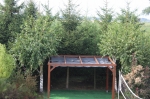 Uitschuifbaar HDPE schaduwdoek op maat gemonteerd op een houten pergola in een tuin omringd door dichte bomen.
