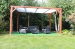 Houten pergola met een wit schaduwdoek van mesh-materiaal, waaronder een set tuinmeubelen op een groene ondergrond.