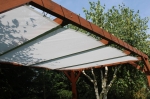 Wit uittrekbaar schaduwdoek van mesh-stof op maat, gemonteerd op een houten pergola in de tuin.