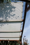 Velă solară mesh albă economică la comandă, montată pe o pergolă din lemn cu umbră de la copaci.