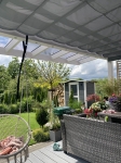 Toldo de sombreamento cinzento sob uma cobertura de terraço em policarbonato, montado sobre móveis de jardim e plantas.