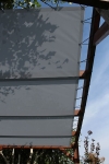 Toldo vela de HDPE a medida instalado en una pérgola de madera con sombras de árboles sobre la tela gris.