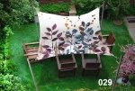 Square 3x3m sun sail with a leaf motif, mounted over wooden garden furniture on a lawn.