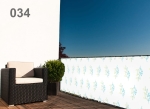 Witte balkonafscherming op maat met groene en blauwe blaadjes, gemonteerd aan een terrasreling met een donkere fauteuil.